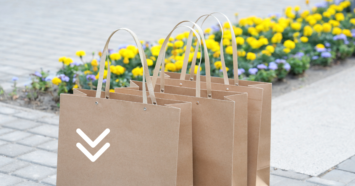 Four plain brown paper shopping bags stand in a row on a paved surface, with a line of yellow and purple flowers in the background.